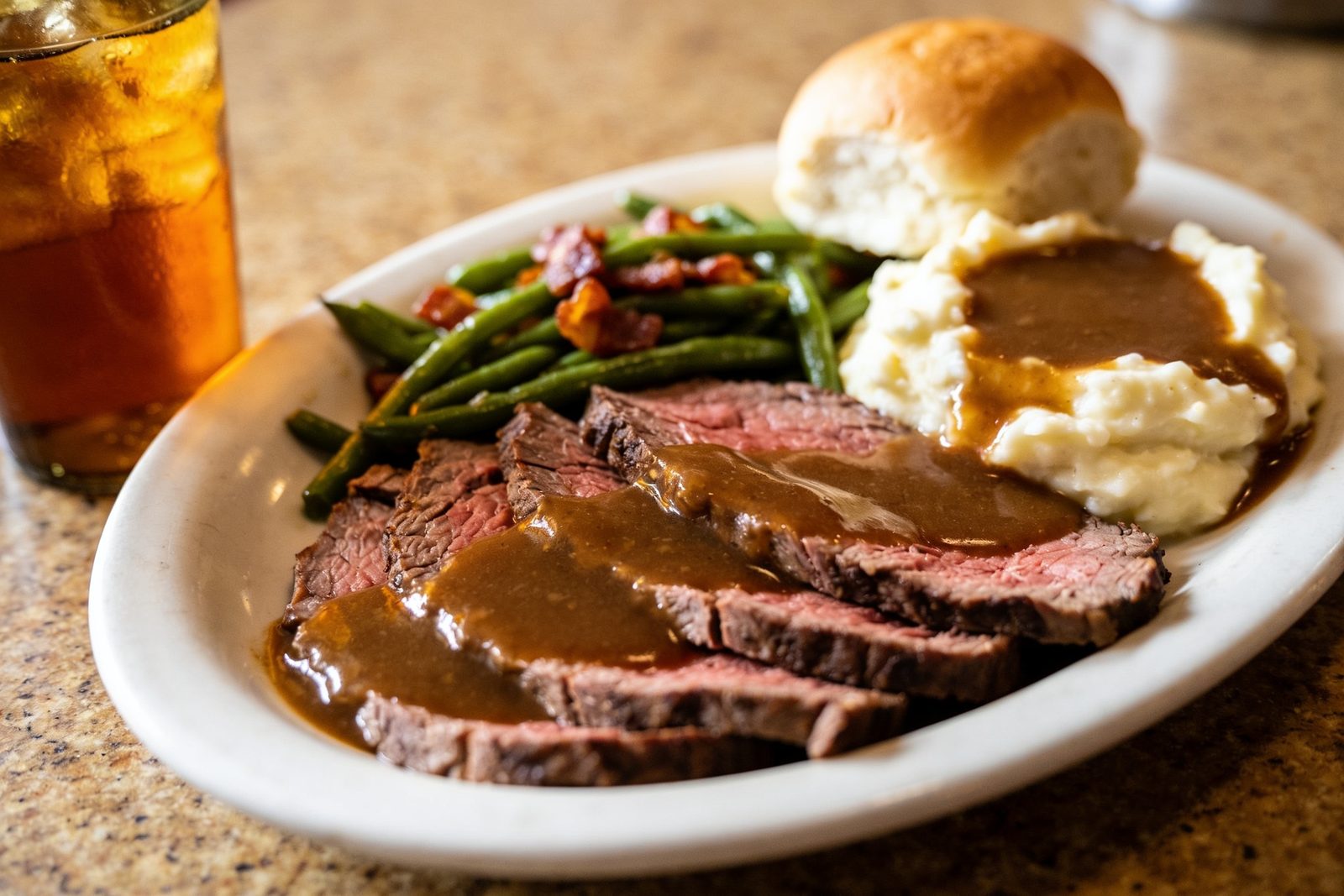 The daily plate lunch: sliced roast beef with mashed potatoes and gravy, green beans and a dinner roll on a heavy white plate.