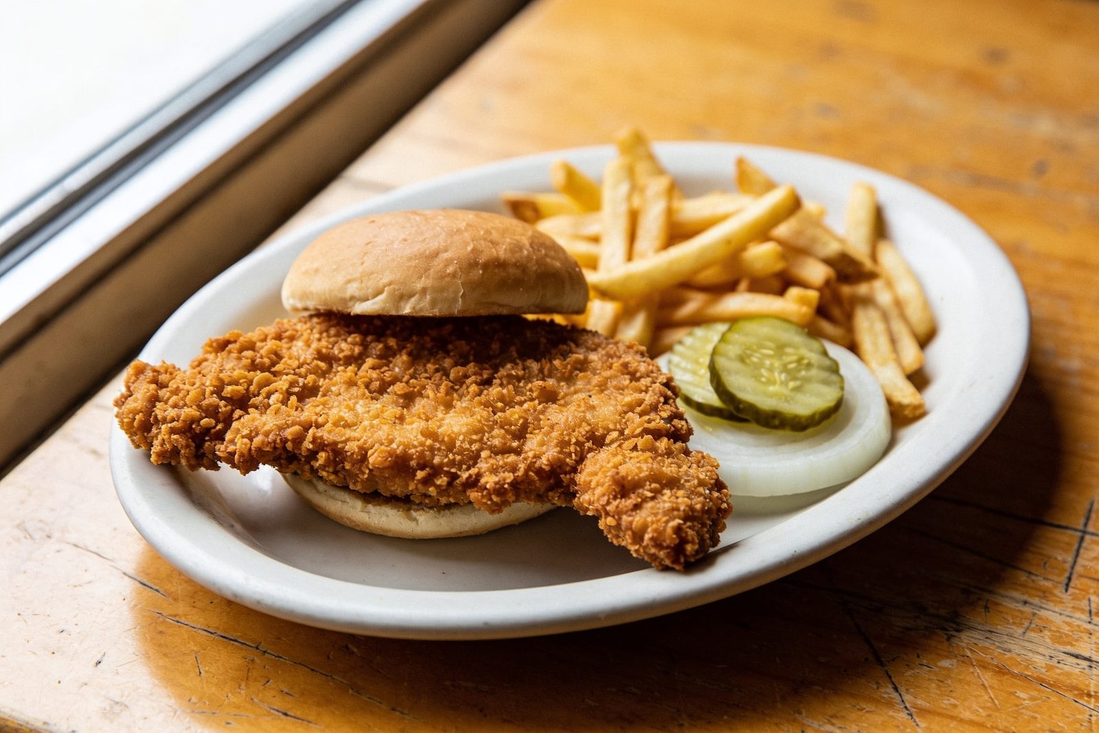A hand-breaded pork tenderloin sandwich on a white oval plate, the golden cutlet hanging well past the bun on every side, pickle chips and raw onion beside it.